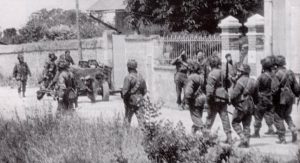 Faldskærmssoldater (med hjelm) og Commandosoldater (med baret) i Amfreville - sandsynligvis kirken i baggrunden))
